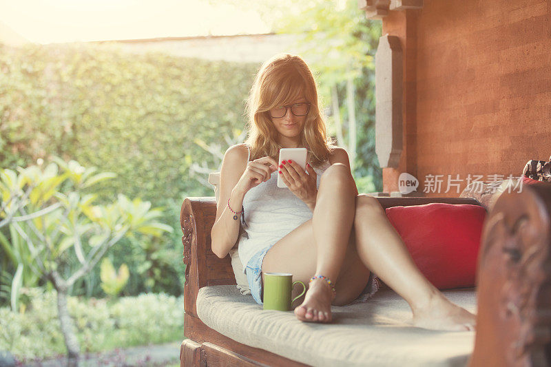 女孩使用手机和喝咖啡/茶在花园的沙发上。